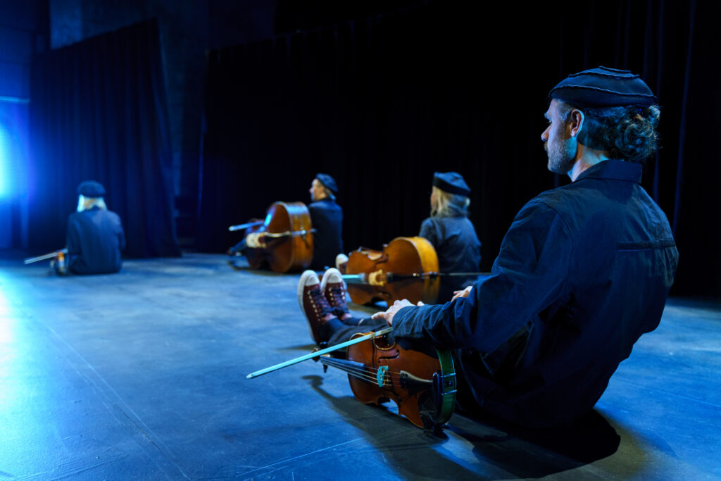 Fyra stråkmusiker iklädda svarta dräkter sitter i ett blått sken på en scen med instrumenten bredvid sig.