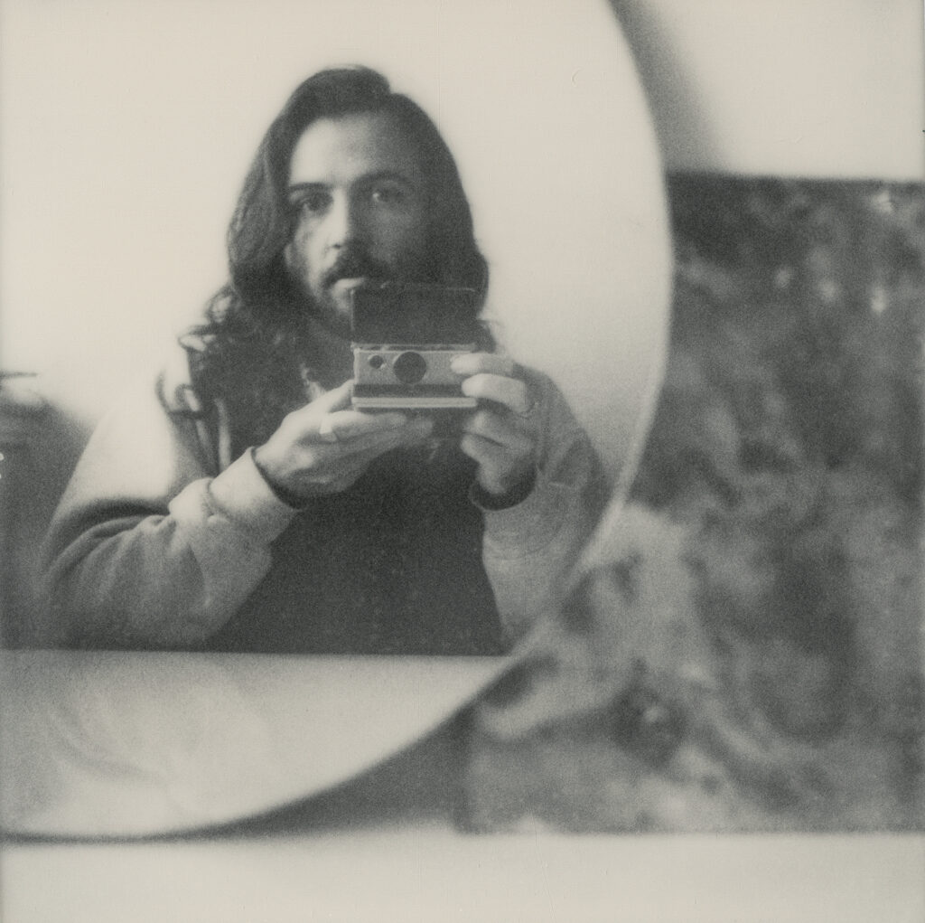 Spegelbilden av en ung man i skägg som håller en kamera i handen.