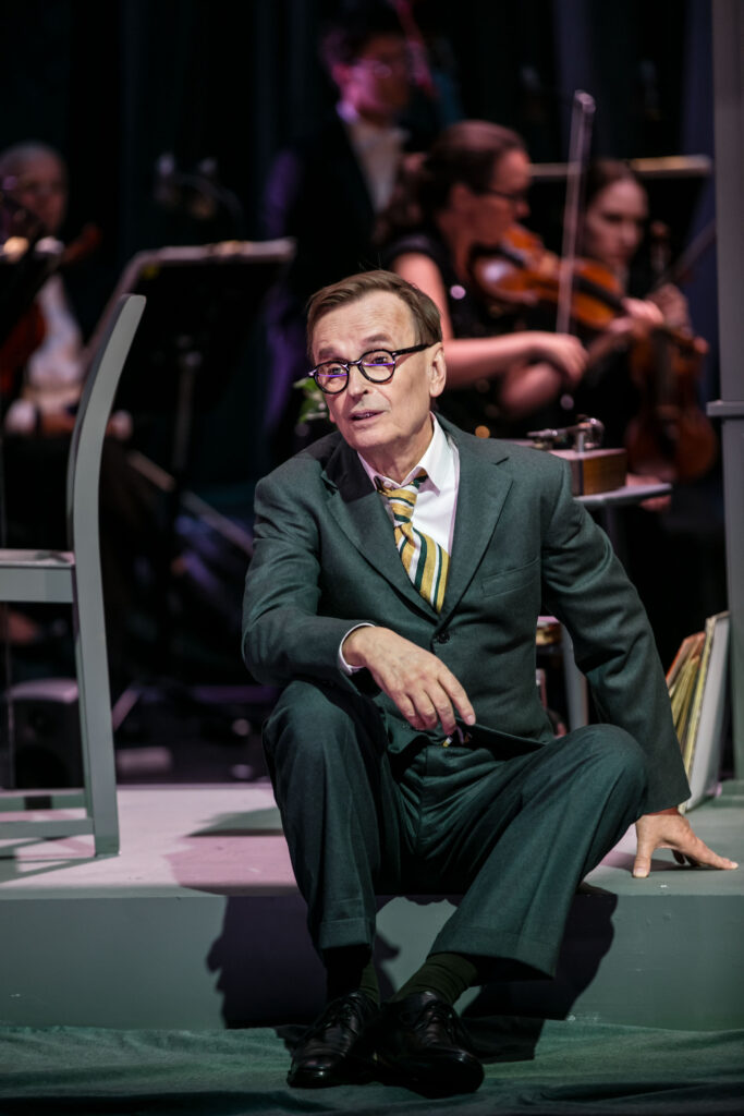 En äldre man i grå kostym och glasögon sitter på en scen och pratar. En orkester spelar i bakgrunden.